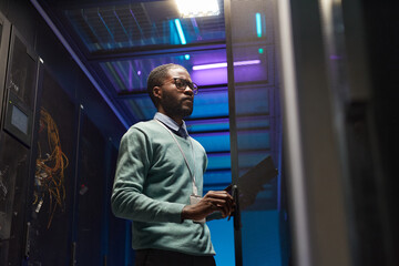 Low angle view at African American data engineer working with supercomputer in server room lit by...