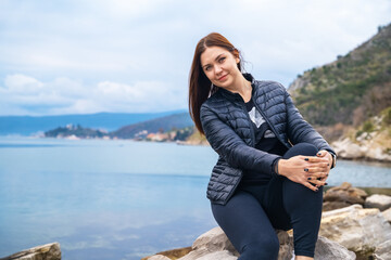 Naklejka na ściany i meble Smiling young attractive woman posing outdoor against mountains and and blue lake or sea.