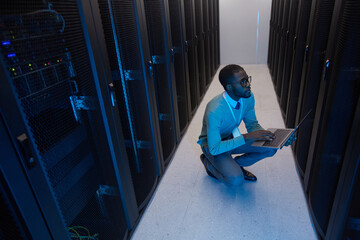 High angle view at young African American data engineer working with supercomputer in server room lit by blue light, copy space