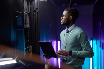Side view portrait of African American data engineer holding laptop while working with supercomputer in server room lit by blue light, copy space