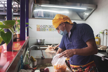 Un hombre con pañoleta amarilla y tapabocas en una cocina abriendo una bolsa con alimentos y a su...