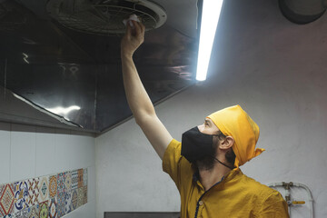 Un hombre con tapabocas y pañoleta amarilla limpiando la ventilación de la cocina con papel