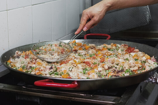 Una olla grande con arroz, tomate, pulpo, carne, camarones, habichuela, alverja y zanahoria picados sobre un fogón en una cocina y la mano de un hombre con una espumadera sobre la preparación