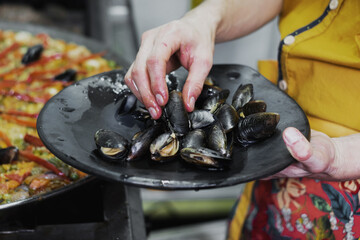 Las manos mojadas de un hombre poniendo mejillones de un plato de barro a una paella dentro de la...