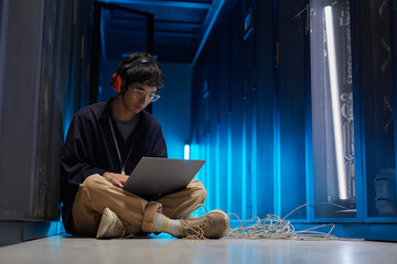Full length portrait of young Asian man sitting on floor in server room lit by blue light while setting up supercomputer network via laptop, copy space