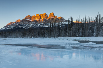Calm Canadian Rockies winter landscape at sunset of a cold river with snow, ice, trees and orange mountains reflected in the clear water, Castle Mountain, Banff National Park, Alberta, Canada
