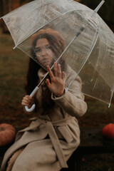 The girl sits on a bench, her hand touches the edge of the umbrella and looks through the transparent umbrella.