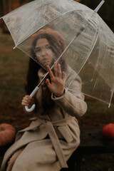 Fototapeta premium The girl sits on a bench, her hand touches the edge of the umbrella and looks through the transparent umbrella.