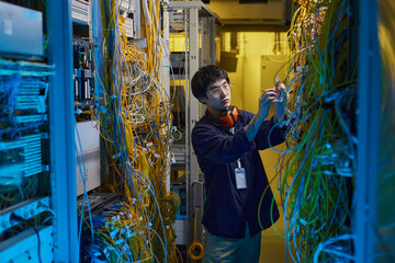 Wide angle portrait of young network technician connecting wires and cables in server room lit by blue light, copy space