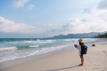 Rear view of a female traveler using and working on laptop computer while walking on the beach