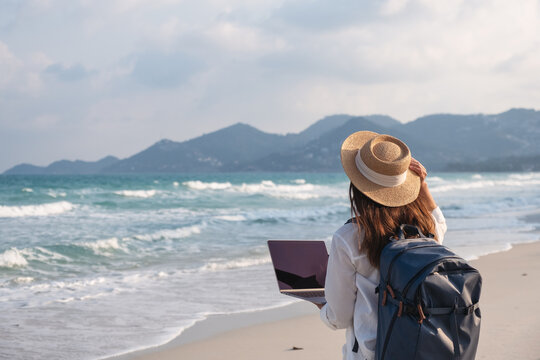 Rear view of a female traveler using and working on laptop computer while walking on the beach