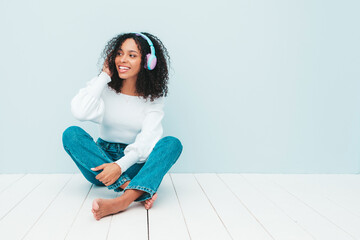 Beautiful black woman with afro curls hairstyle.Smiling model in sweater and jeans.Sexy carefree female listening music in wireless headphones.Sitting in studio near light blue wall