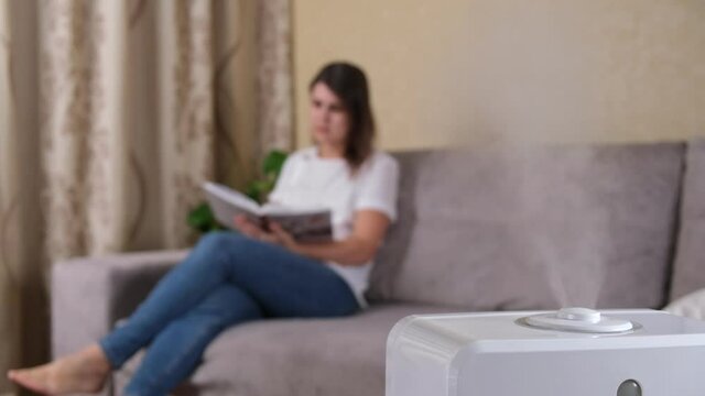 Steam from a working humidifier with ionization of air in the room, against a blurred background, woman reading a book sitting on the couch