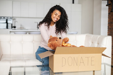 Kind smiling African American female volunteer with curly hair sitting on a sofa, packing toys in a...