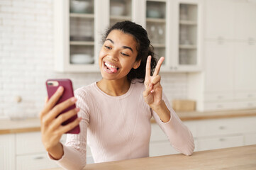 Funny biracial teen with dark hair and bronze skin showing tongue and taking photo on the phone, making a selfie, showing a piece sign, at home in the kitchen by the tabletop, chatting with friends