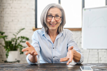 Happy friendly senior female teacher manager with pleasant smile in earphones and glasses looking...