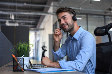 Cheerful young support phone male operator in headset, at workplace while using laptop, help...