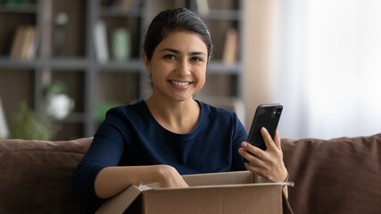 Portrait of smiling young Indian woman unpack box parcel with internet order shopping online on smartphone. Happy ethnic female buyer excited with good quality delivery service. Shipping concept.