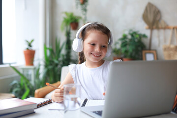 Smiling little girl in headphones handwrite study online using laptop at home, cute happy small child in earphones take Internet web lesson or class on PC.