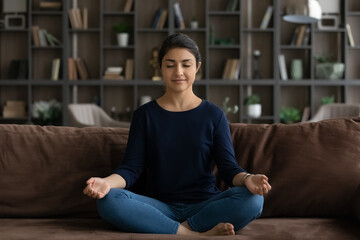 Calm millennial Indian woman sit on sofa in living room practice yoga with mudra hands. Young mixed...