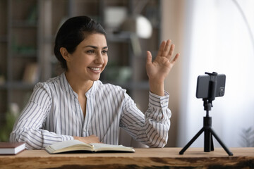Happy Indian female teacher or tutor wave greet talking on video call on smartphone. Smiling young...