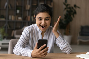 Overjoyed millennial Indian woman look at cellphone screen triumph win lottery online. Excited...