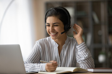 Overjoyed millennial Indian female student in headphones look at laptop screen study online from home. Happy young mixed race woman in earphones talk on video call with teacher. Virtual event concept.