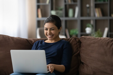 Naklejka premium Smiling millennial Indian woman relax on couch in living room look at laptop screen talk on video call. Happy young mixed race female use computer watch movie, have webcam online virtual conference.