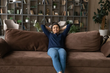 Happy millennial Indian woman sit relax on comfortable couch in living room sleep or take nap. Calm young mixed race female renter rest on sofa at home, daydream enjoy leisure weekend. Rent concept.