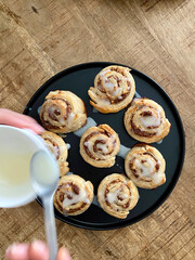 Homemade Cinnamon Rolls or Cinnamon Buns Pouring Cream Sauce on Wooden Surface.