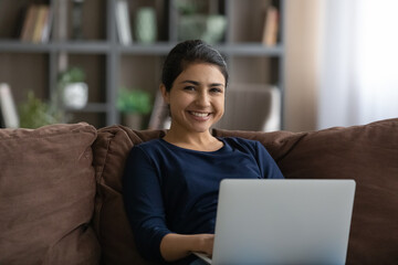 Portrait of smiling millennial Indian woman sit rest on sofa in living room use modern laptop gadget. Happy young mixed race female work online on computer at home office. Technology user concept.
