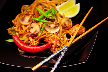 Stir-fried noodles on black plate with mushrooms and chicken meat, and food sticks
