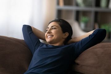 Fototapeta premium Smiling young Indian woman lying resting on comfortable couch in living room look in distance dreaming or thinking. Happy millennial ethnic female relax on sofa at home. Peace, stress free concept.