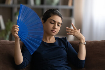 Unwell young Indian woman sit rest on sofa at home feel overheated wave with hand fan. Unhealthy...