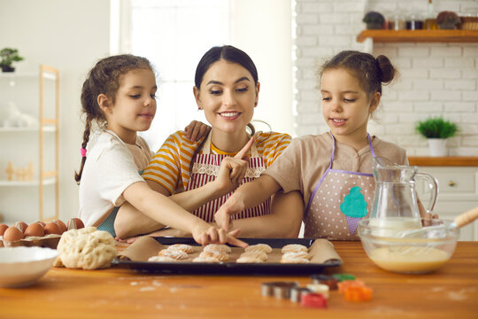 Happy mother baking pastries with children. Young woman teaching daughters to make biscuits. Cute impatient kids can't wait for sweet cookies to be ready while cooking together with mom in the kitchen