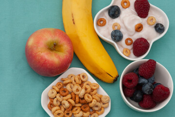 Breakfast - strawberry yogurt with cereal rings, raspberries and blueberries, pink apple and non-pealed banana, top view, on the light blue background