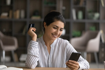 Smiling young Indian woman shopping online on smartphone with credit card. Happy millennial mixed race female buyer client feel excited buy on internet using secure banking system on cellphone.