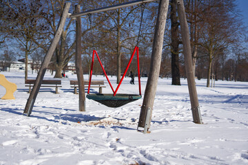 Large swing on a playground in the park, trees and building in the background. red dominates. Blue sky. Winter with snow in germany.