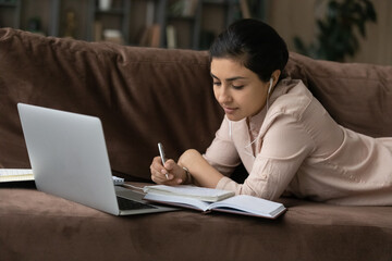 Focused millennial Indian female student lying on sofa in living room study online using computer. Concentrated mixed race woman busy working or taking distant course on laptop. Education concept.