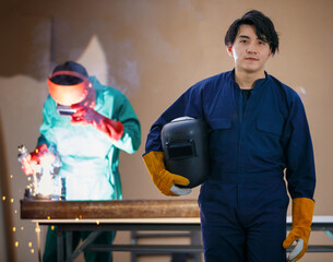 young confident positive Asian man wearing a Blue factory uniform holding a welding mask standing...