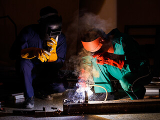 two engineers working in the dark at nighttime. Mechanics wearing a mechanic coveralls work together to weld the metal rod, making nice spark, bokeh, and smoke