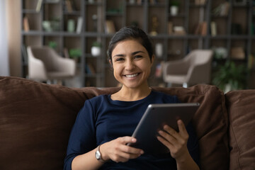 Fototapeta na wymiar Portrait of smiling young Indian woman sit relax on sofa in living room use tablet gadget browsing wireless internet. Happy mixed race female rest at home with modern pad device text message online.