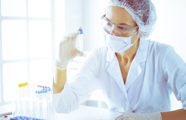 Professional female scientist in protective eyeglasses researching tube with reagents in sunny laboratory toned in blue. Medicine and science researching