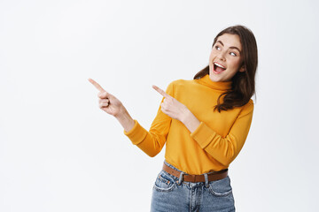 Cheerful young female model smiling, pointing fingers left at logo with amused face, checking out...