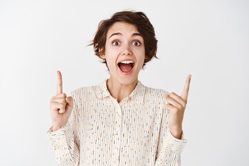 Surprised woman scream of excitement and joy, pointing fingers up, showing advertisement on white...
