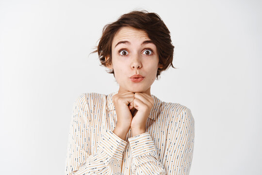 Intrigued young woman looking with interest and surprised at camera, raising eyebrows and gazing silly, standing in blouse over white background