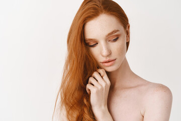 Beauty and skincare. Young woman with natural long red hair and pale skin, looking down, posing naked over white background