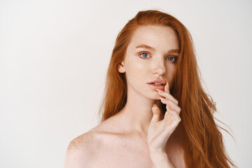 Close-up of gorgeous redhead lady with pale smooth skin and no makeup, touching lips and looking at camera thoughtful, standing naked on white background