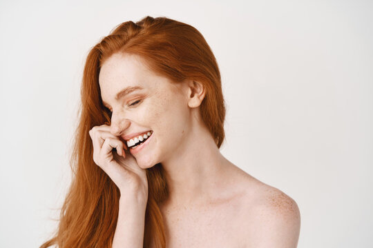 Beauty. Close-up of happy redhead woman with pale perfect skin, laughing and showing white teeth, standing naked on studio background
