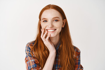 Fototapeta premium Close-up of redhead female without make-up and white perfect smile, looking happy at camera, standing against white background